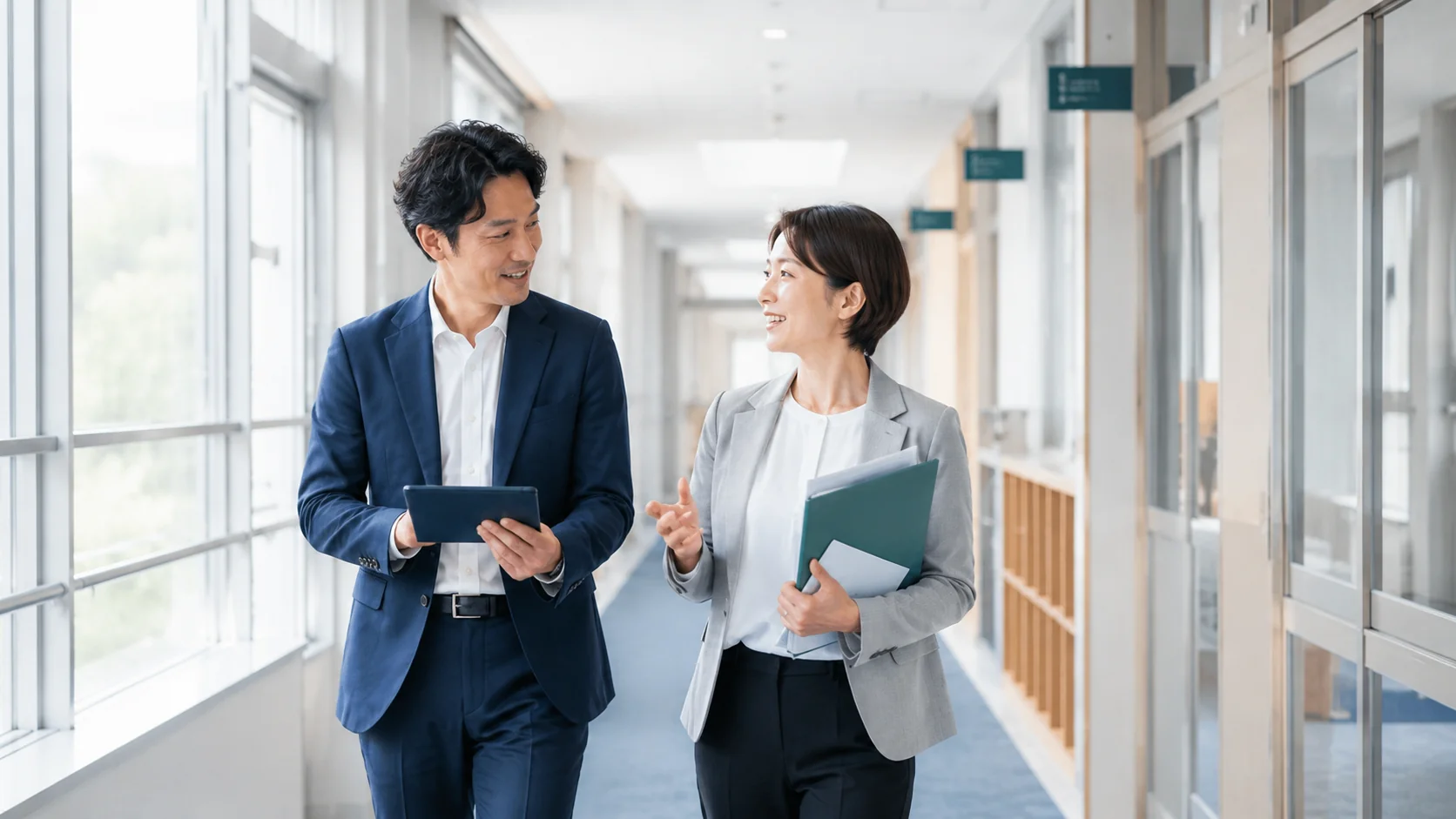 自然光の入る校舎の廊下で、タブレットを手に対話しながら歩く2人のイメージ